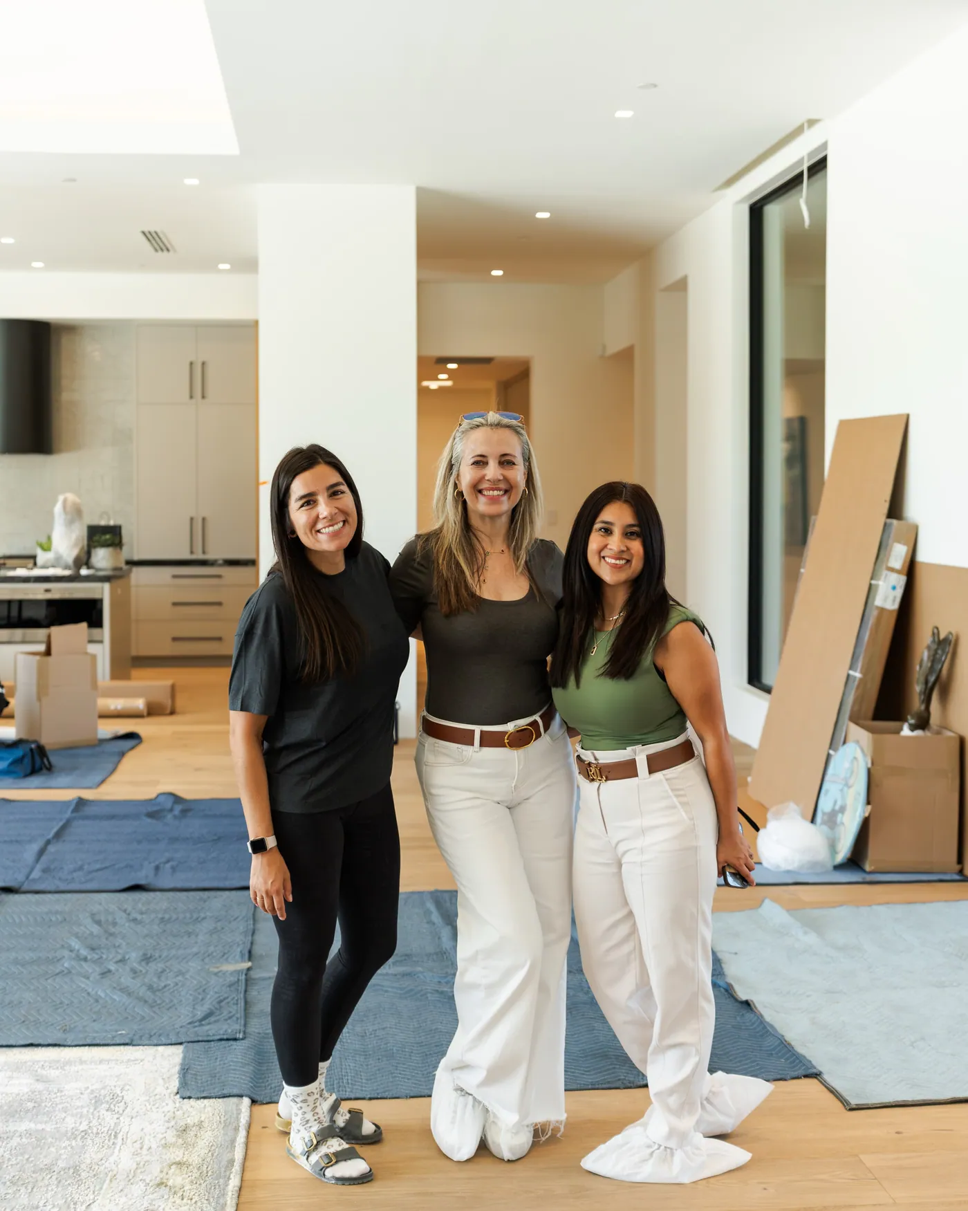Interior design installation coordinated with Arizona luxury home builders: three members of the PHX Interior Design team smiling in a newly finished luxury home on installation day, with shoe covers on and protective blankets over the oak floors