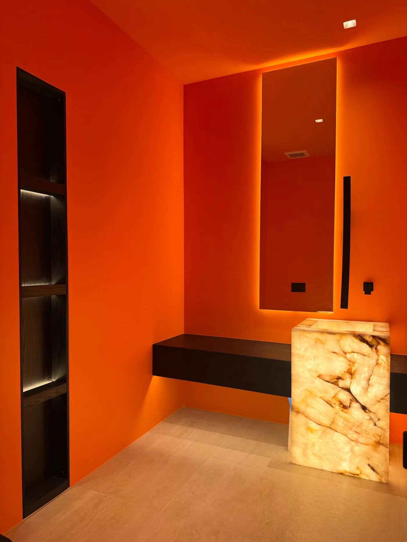 Backlit onyx feature powder room for design-build luxury homes: powder room in saturated orange with a backlit onyx pedestal sink, a floating dark wood shelf, an LED-lit mirror and a recessed illuminated niche