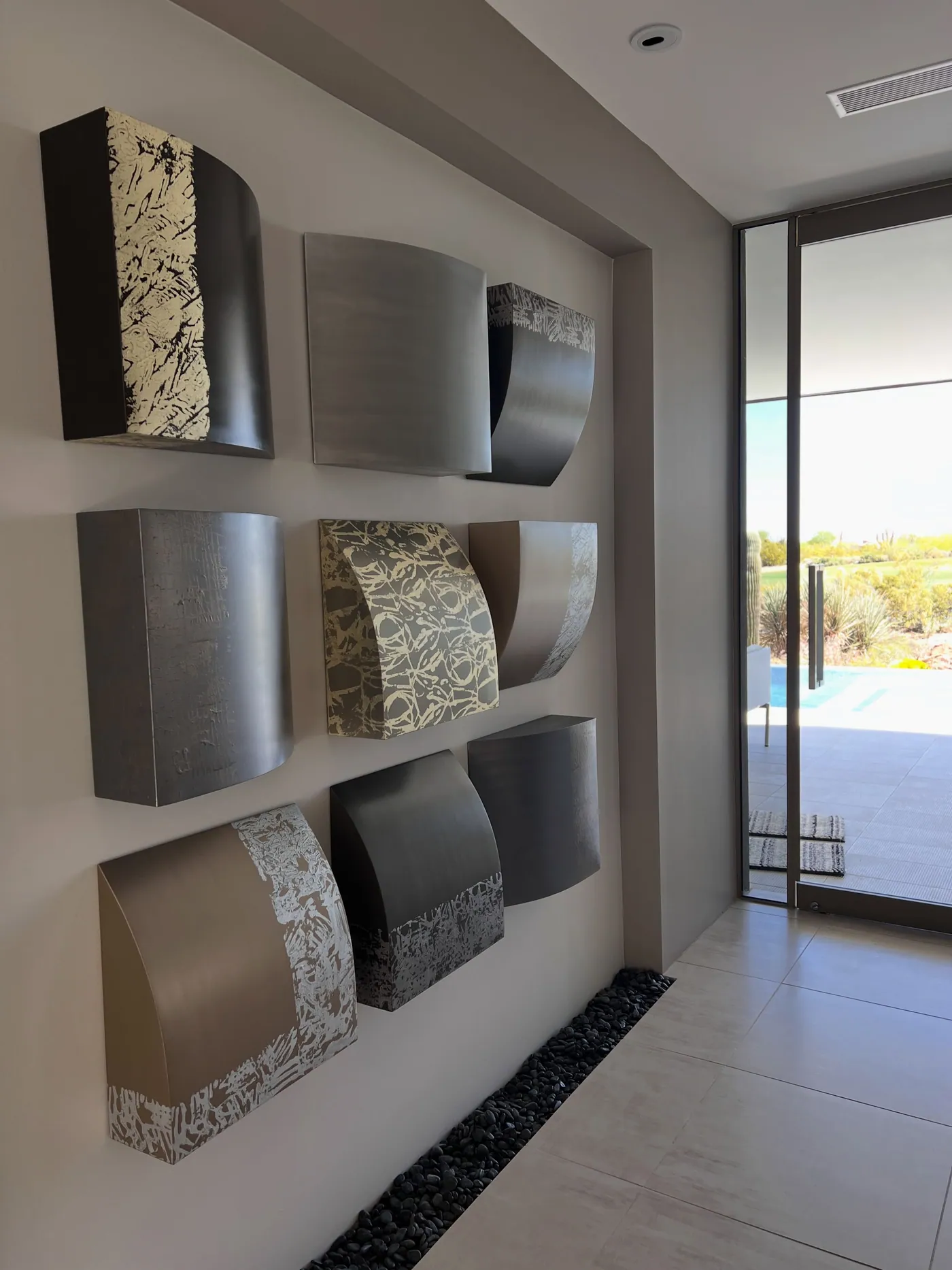 Custom art wall and millwork coordination for design-build custom homes: wall of nine sculptural curved metal panels in bronze, pewter and gold leaf in a luxury home entry, with a glass pivot door opening to a golf course and pool