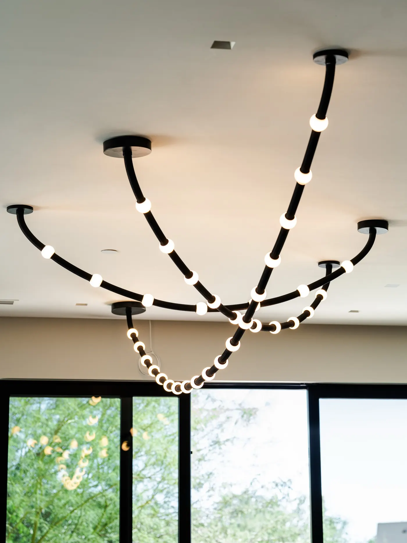 Custom lighting design by a Scottsdale luxury interior designer | PHX Interior Design Custom lighting design by a Scottsdale luxury interior designer: custom black-cord ceiling light with glowing spherical globes suspended from multiple canopies in front of tall windows and a palo verde tree
